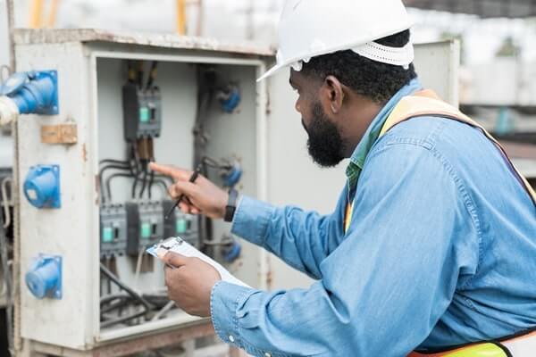 An electrician in a blue shirt and white hard hat checks the voltage inside an electrical box
