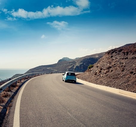 Thumbnail of a small car driving along a curved mountain road under a blue sky next to the sea