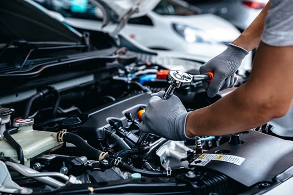 A mechanic wearing gloves uses a wrench to make an adjustment under the hood of a car