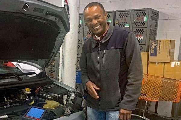 Nigel Campbell, instructor in the Automotive Service & Repair program smiles next to a car with the hood up