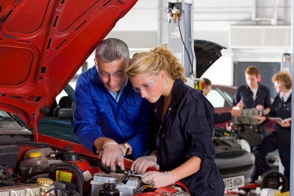 An instructor and a student work under the hood of a car in an automotive shop