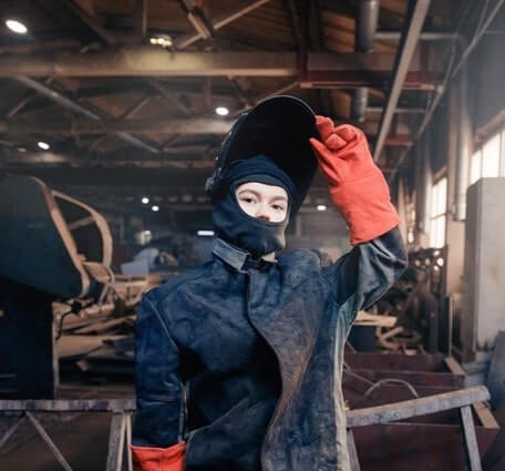 Thumbnail of a welder stands in the shop wearing red gloves and lifting her protective face mask